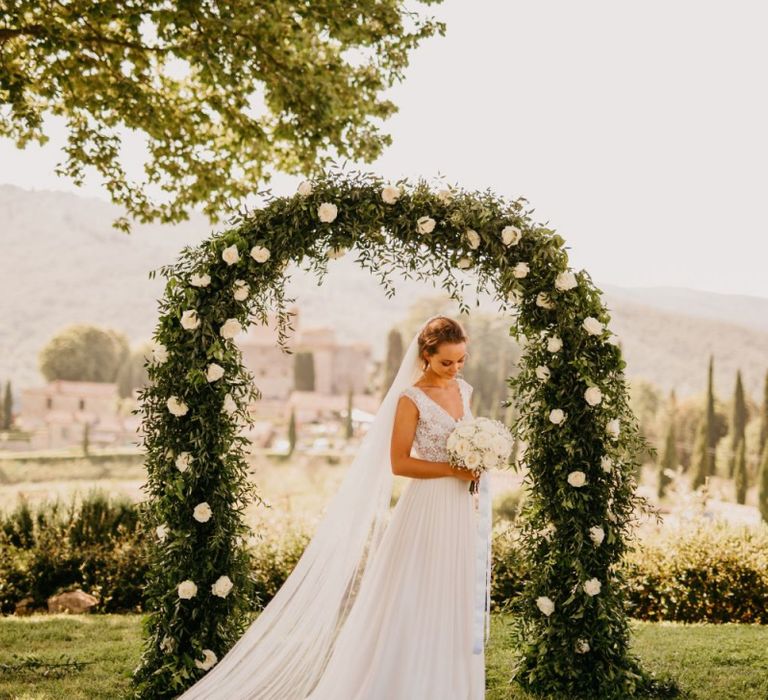 Bride in Bespoke Wedding Dress Standing in Front of White and Green Floral Arch