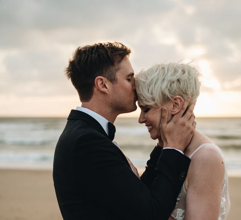 Groom in Tuxedo Kissing His Brides Forehead on the Beach