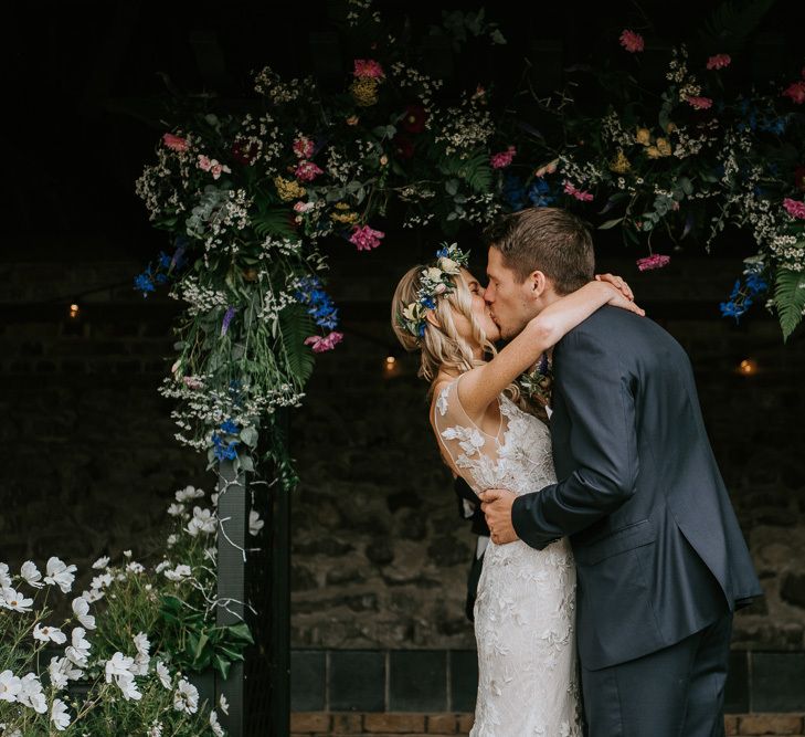 Bride in Lace Watters Wedding Dress and Groom in Navy Hugo Boss Suit Kissing at the Flower Covered Altar