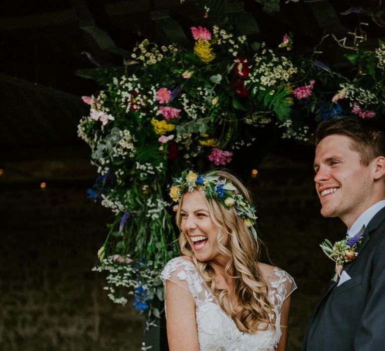 Bride in Lace Watters Wedding Dress and Groom in Navy Hugo Boss Suit Holding Hands Laughing at the Flower Covered Altar