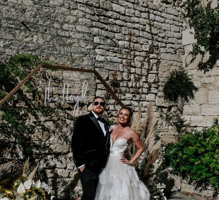 Stylish Bride in Feather Skirt Wedding Dress and Groom in Black tie Suit Standing Next to a Hexagon Backdrop