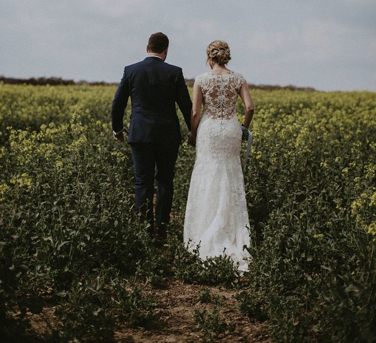 Bride in Lace Back Maggie Sottero Wedding Dress and Groom in Navy Suit Walking Through Fields