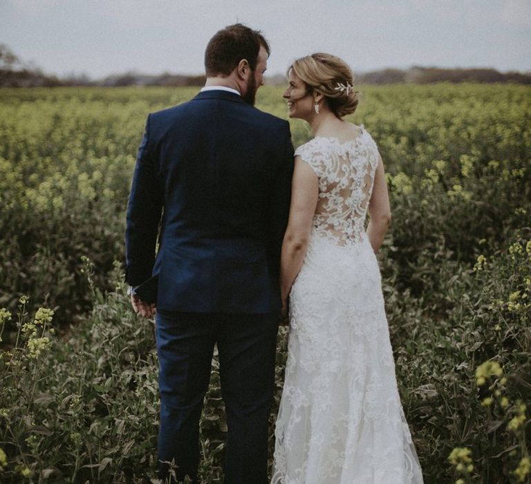 Bride in Lace Back Maggie Sottero Wedding Dress and Groom in Navy Suit Holding Hands in a Field