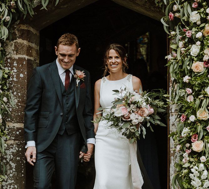 Bride in Mikaella Bridal Wedding Dress and Groom in Three-piece Clifton Suit Exiting The Church Under a Floral Arch