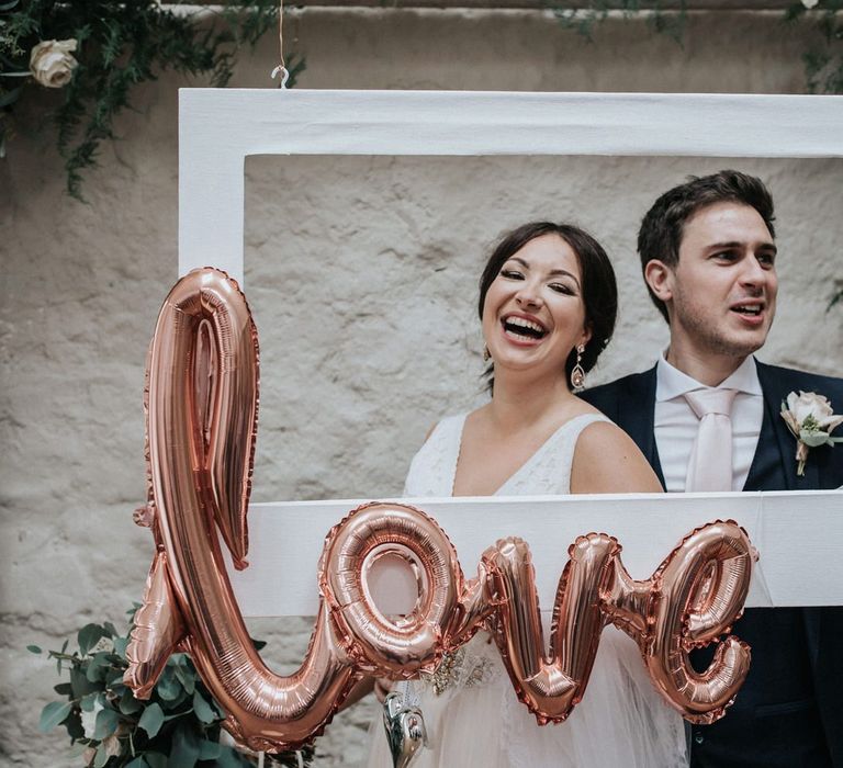 Bride and groom posing at Selfie Station with white frame and gold foil balloon.