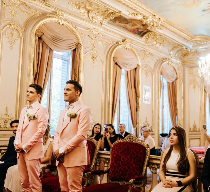 Grooms wearing pink wedding suits in ornate and golden ceremony room in Paris city hall