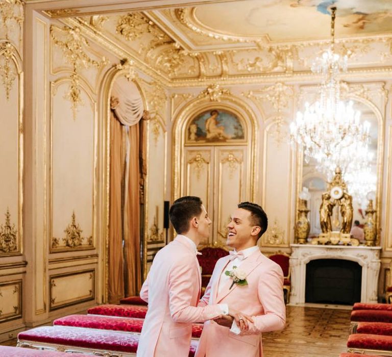 Grooms holding hands and smiling in grand room in Paris with gold and ornate styling