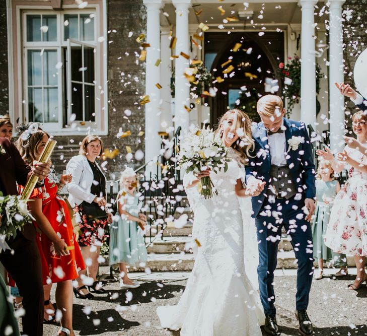 Confetti Moment | Bride in Pronovias Oringo Lace Bridal Gown &amp; Cape | Groom in Blue Marks and Spencer Suit | Green, White &amp; Gold Wedding at Buckland Tout Saints, Devon |  Darina Stoda Photography