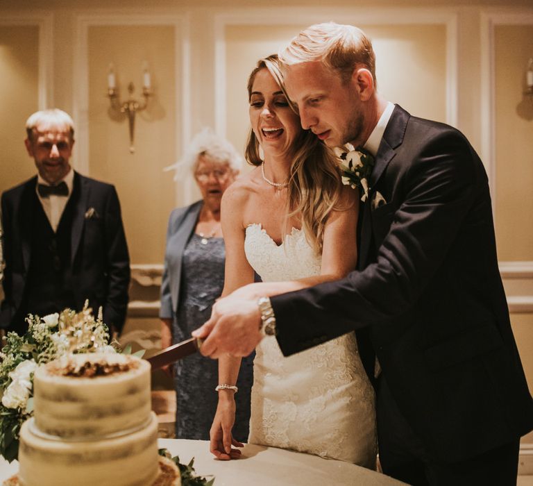 Cutting the Wedding Cake | Bride in Pronovias Oringo Lace Bridal Gown &amp; Cape | Groom in Blue Marks and Spencer Suit | Green, White &amp; Gold Wedding at Buckland Tout Saints, Devon |  Darina Stoda Photography