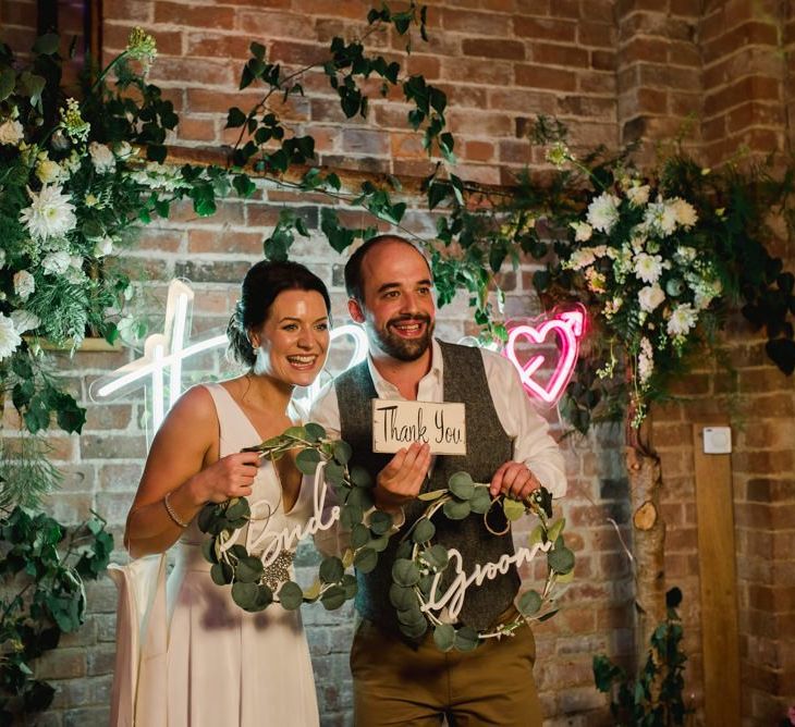 Bride and groom pose with personalised neon sign and white wedding flowers hoop decor