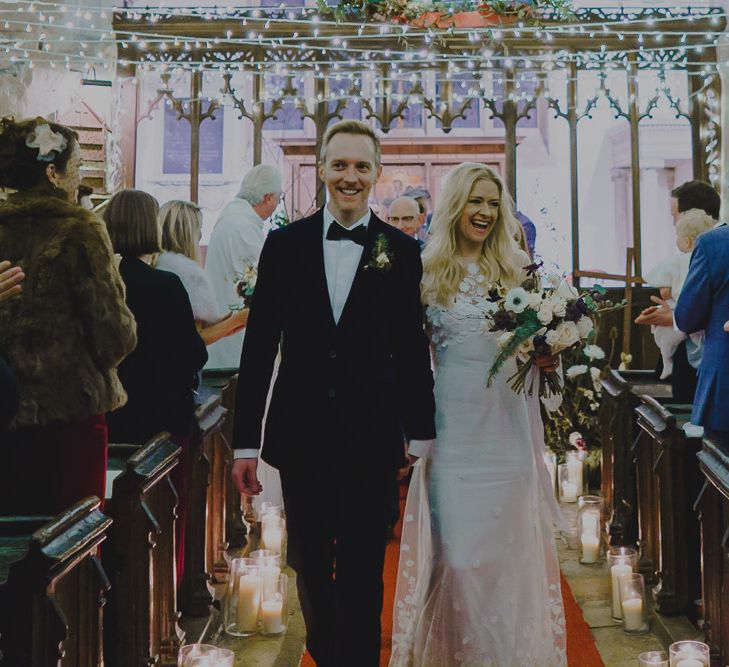 Bride in lace dress and groom in velvet dinner jacket walking up the church aisle as husband and wife