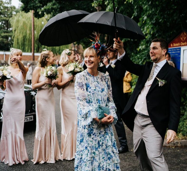 Mother of the bride and bridesmaids stand, smiling, underneath umbrellas as they wait for the bride to exit her car