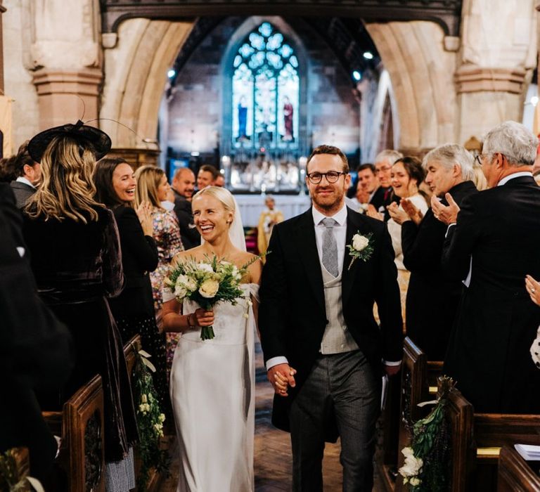 Bride and groom hold hands and walk back down the aisle as husband and wife while smiling at their guests ]