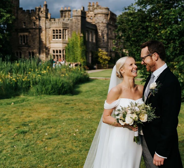 Bride in off the shoulder wedding dress and groom in traditional morning suit standing in front of Hawarden Castle in Wales