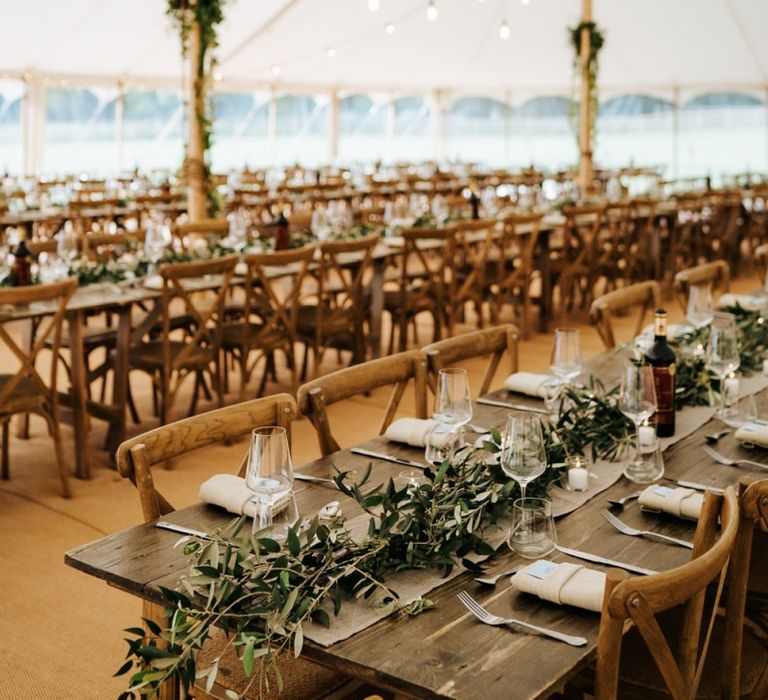 Marquee reception with wooden tables, greenery table runners and hanging festoon lights