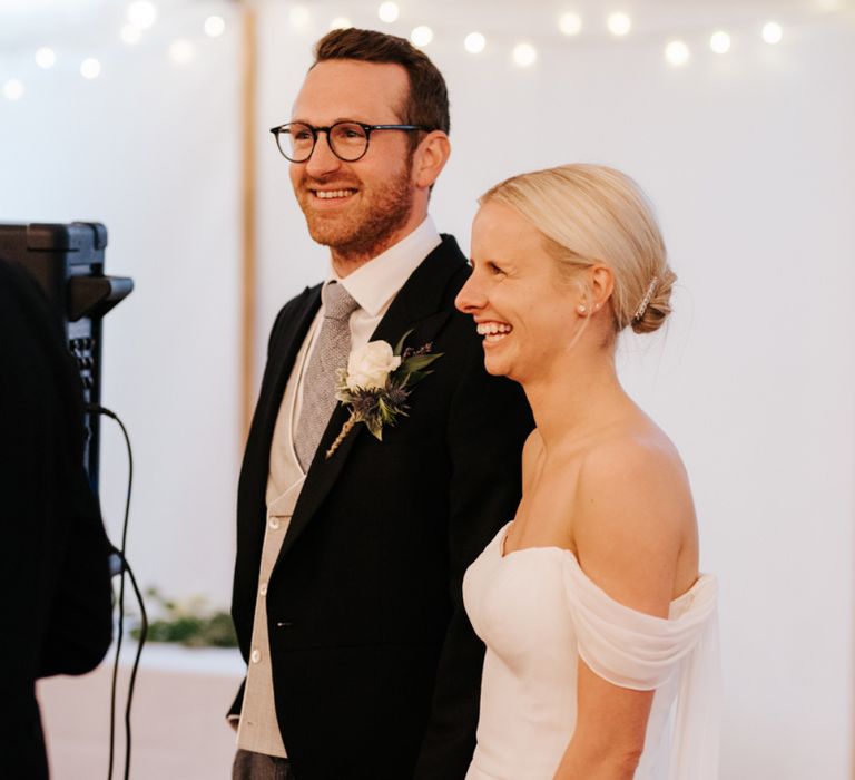 Bride and groom laugh and smile, holding hands, while fathe of the bride delivers wedding speech