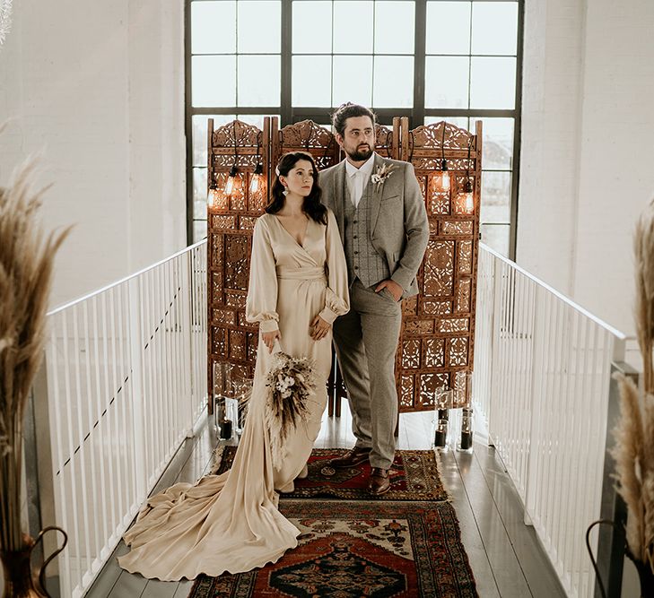 Bride in Satin Wedding Dress and Groom in Wool Suit with Top Knot Standing in Front of a Moroccan Screen and Rug