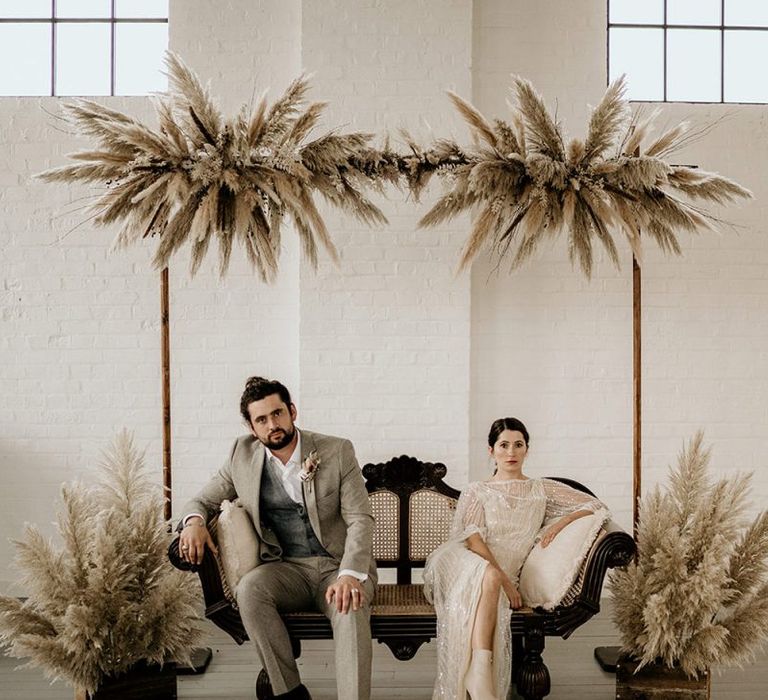 Bride in Sequin Wedding Dress and Groom in Wool Suit with Top Knot Sitting on a Sofa under a Pampas Grass installation