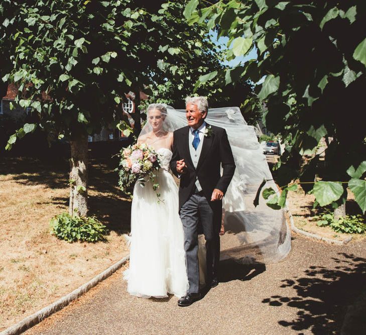 Bride in Tulle and Applique Halfpenny London Wedding Dress and Father of The Groom Walking Through the Church Courtyard