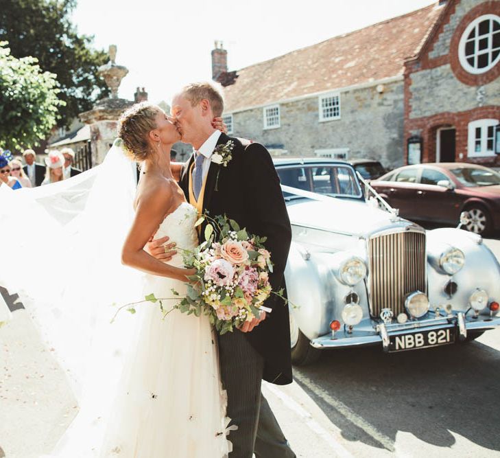 Bride in Halfpenny London Wedding Dress  with Braided Up Do and Groom in Traditional Morning Suit Kissing Next to Their Wedding Car