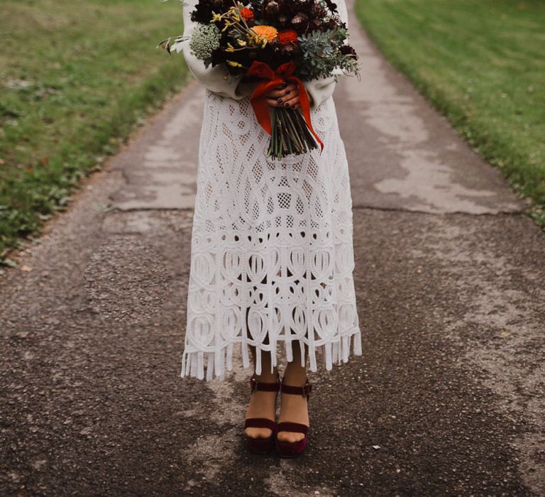 Bride in Crochet Wedding Dress and Woollen Jumper Holding a Orange and Red Dahlia Wedding Bouquet