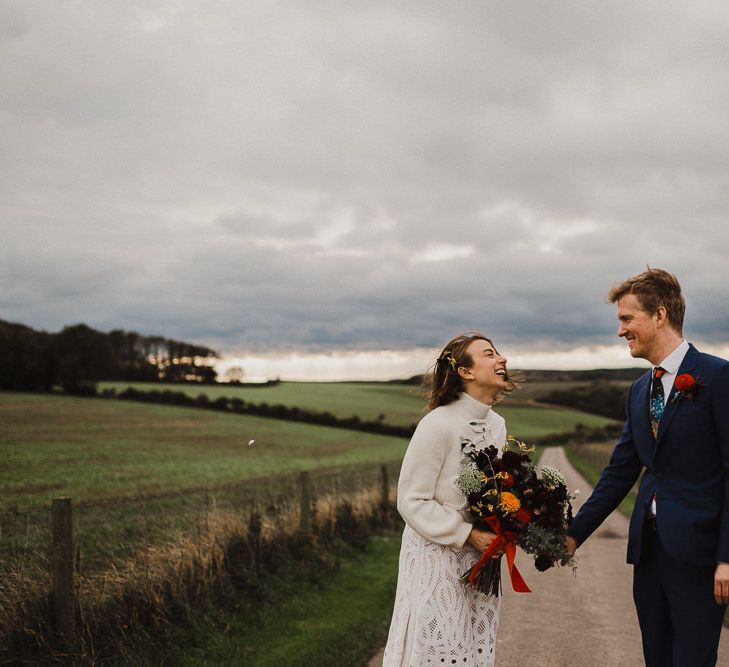 Bride in Crochet Wedding Dress and Woollen Jumper and Groom in Navy Paul Smith Suit Holding Hands on a Country Road