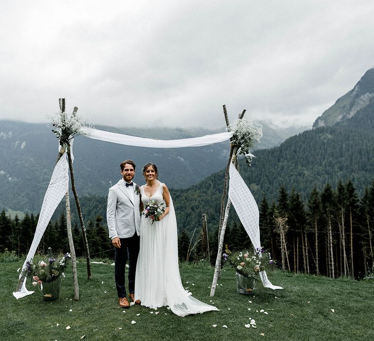 Bride in Mariées Passion Wedding Dress and Groom in Grey Blazer  Standing in Front of Their Draped Wedding Altar