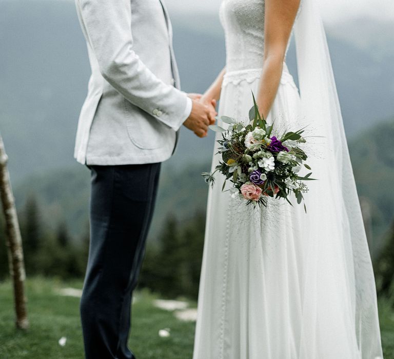 Bride in Mariées Passion Wedding Dress with Pretty Bouquet and Groom in Grey Blazer Holding Hands