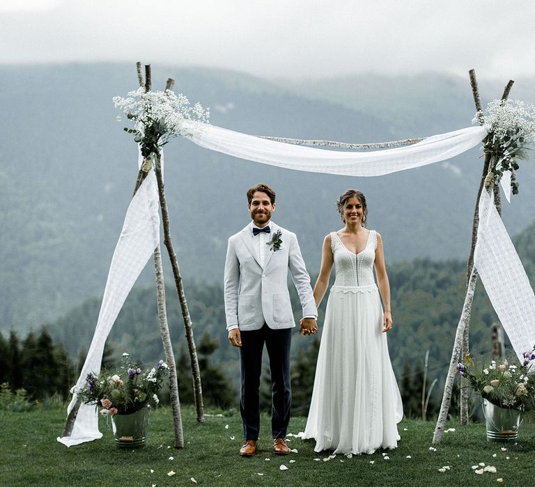 Bride in Mariées Passion Wedding Dress and Groom in Grey Blazer and Navy Chinos Standing in Front of Draped Altar