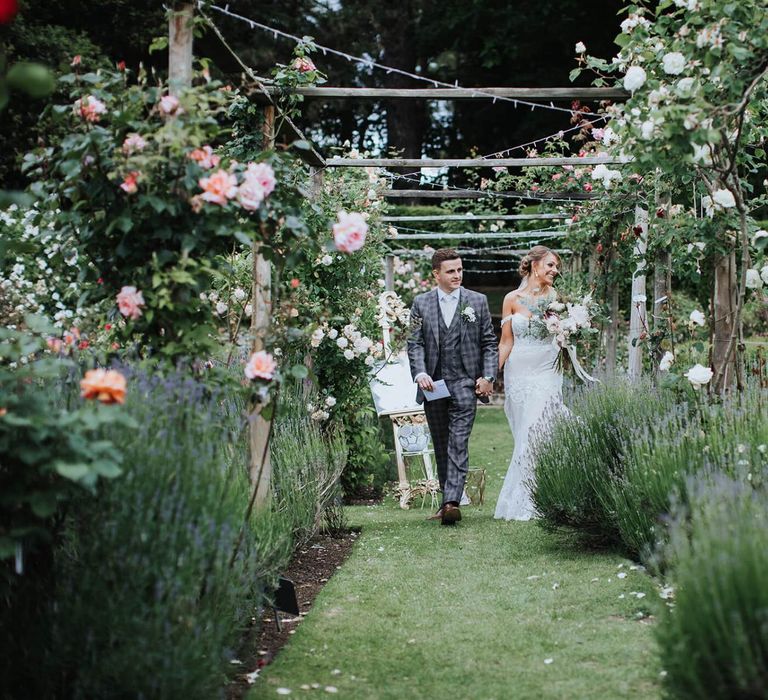 Bride And Groom Walk Through Grounds With Floral Backdrop