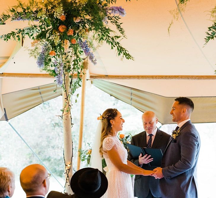 Wedding Ceremony | Exchanging Vows | Bride in Laced KatyaKatya Wedding Dress with Cap Sleeves and Ribbon Belt | Groom in Blue T.M Lewin Suit with Navy Hawkes Bespoke Outfitters Waistcoat | Arch of Flowers | Lace KatyaKatya Dress for Tipi Wedding at Fforest Farm | Claudia Rose Carter Photography