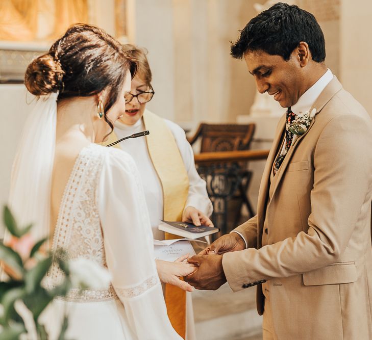 Groom in beige suit putting on his brides wedding ring