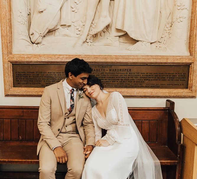 Bride in Rosa Clara wedding dress with long sleeves and groom in beige suit sitting on a wedding pew