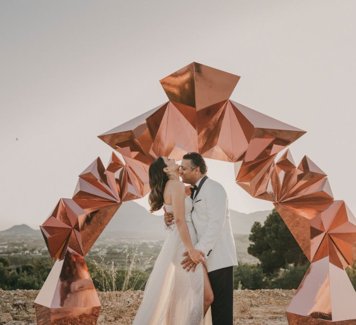 Bride in One Shoulder Muse by Berta Wedding Dress and Groom in White Dinner Jacket Standing in Front of a Copper Geometric Structure