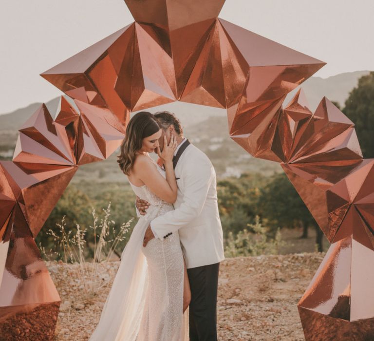 Bride in One Shoulder Muse by Berta Wedding Dress and Groom in White Dinner Jacket Kissing in Front of a Copper Geometric Structure