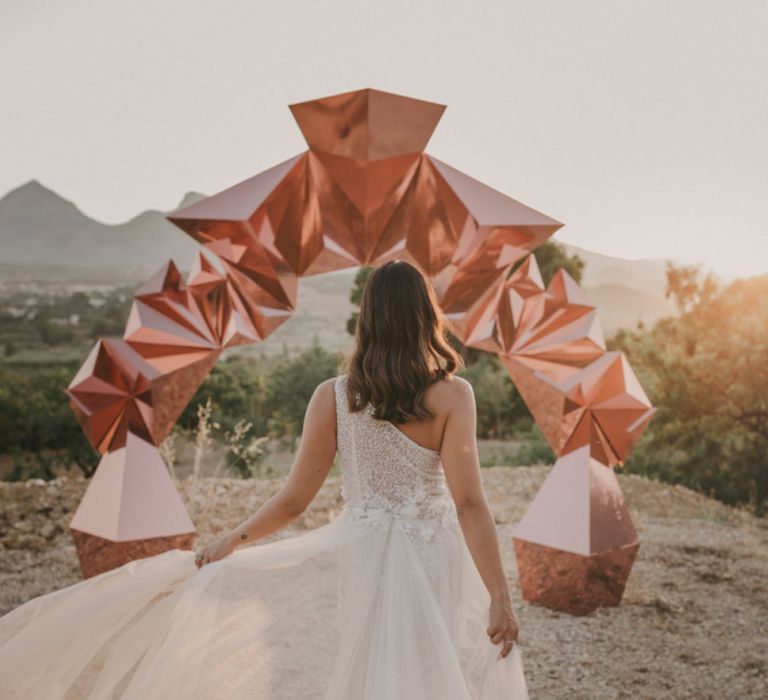 Bride in One Shoulder Muse by Berta Wedding Dress with Tulle Train Standing in Front of a Copper Geometric Structure