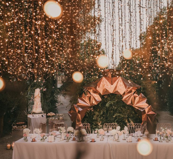 Top Table Wedding Decor with Fairy Light Canopy, Copper Geometric Structure, Flower Stems in Vases and Geode Wedding Cake