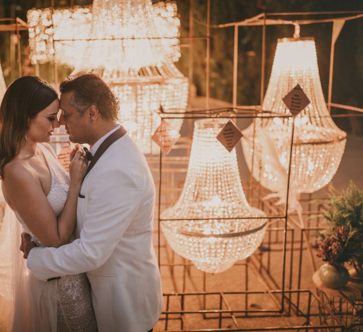 Bride in One Shoulder Wedding Dress and Groom in White Dinner Jacket Standing in Front of Their Chandelier Table Plan