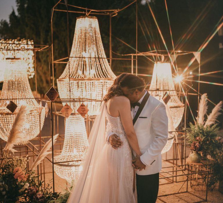 Groom in White Dinner Jacket Embracing his Bride in Front of a Chandelier Table Plan