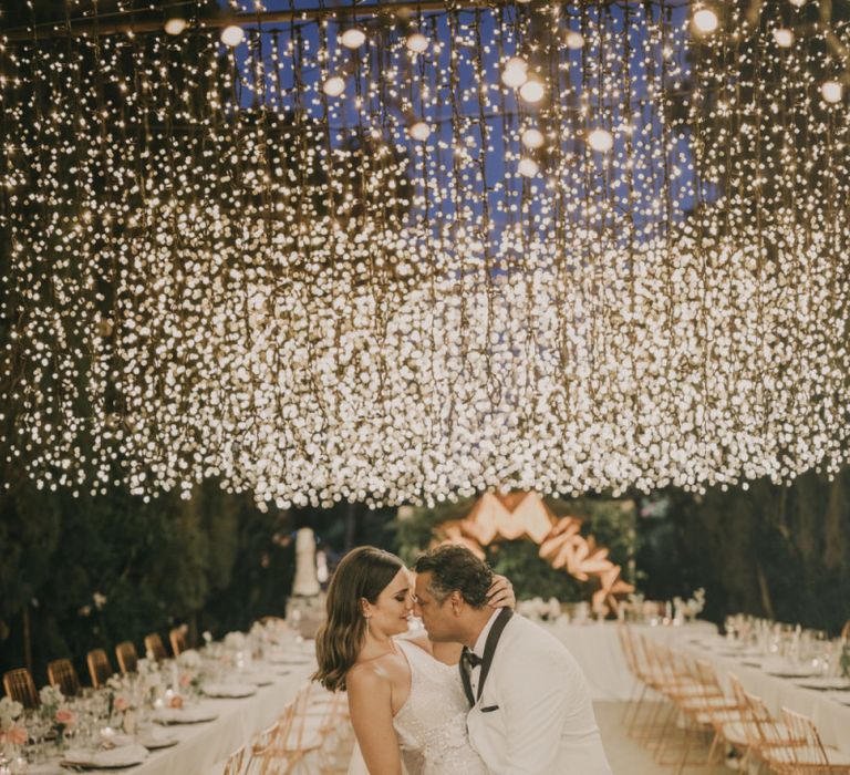 Bride in One Shoulder Muse by Berta Wedding Dress and Groom in White Dinner Jacket Posing Under a Fairy Light Canopy