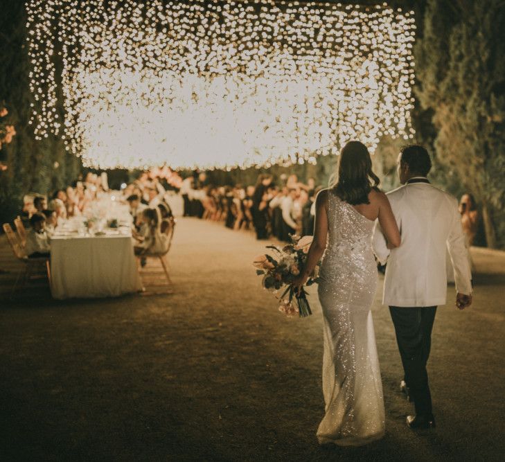 Bride in One Shoulder Muse by Berta Wedding Dress and Groom in White Dinner Jacket Entering the Outdoor Wedding Reception with Fairy Light Canopy