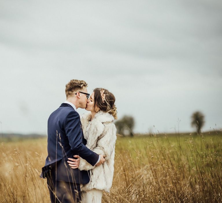 Bride in Faux Fur Coat and Groom in Navy Suit Kissing in the Fields