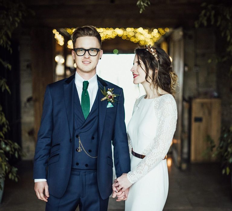 Bride in French By Wendy Makin Wedding Dress and Groom in Navy Blue Suit Holding Hands inside Stone Barn Cotswolds