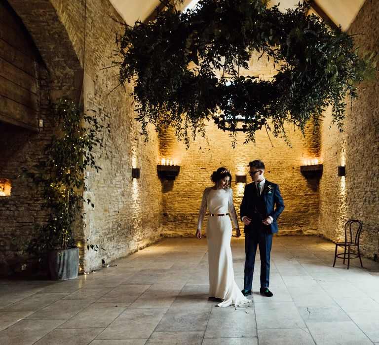 Bride in French By Wendy Makin Wedding Dress and Groom in Navy Blue Suit Holding Hands inside Stone Barn Cotswolds