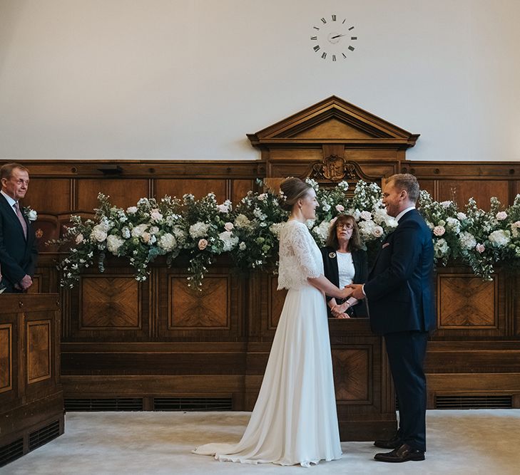 Bride in Charlie Brear Carenne Dress with Spaghetti Straps and Flowing Skirt | Bridal Lace Jacket by Charlie Brear | Groom in Navy Three-Piece Suit with Blush Pocket Square | Blush and White Flowers with Foliage Altar Arrangement | Town Hall Hotel Wedding Ceremony | Statement Floral Arrangement Altar and Potted Plants with Copper Frame Table Plan and Lace Jacket | Miss Gen Photography