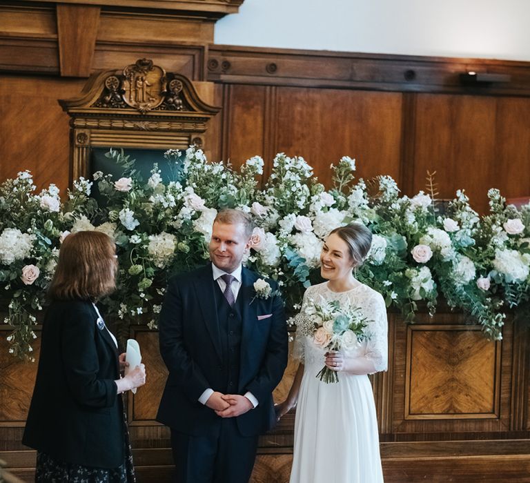 Blush and White Flowers with Foliage Altar Arrangement | Bride in Charlie Brear Carenne Dress with Spaghetti Straps and Flowing Skirt | Bridal Lace Jacket by Charlie Brear | Bouquet of White and Blush Flowers with Foliage | Groom in Navy Three-Piece Suit with Blush Pocket Square | Town Hall Hotel Wedding Ceremony | Statement Floral Arrangement Altar and Potted Plants with Copper Frame Table Plan and Lace Jacket | Miss Gen Photography