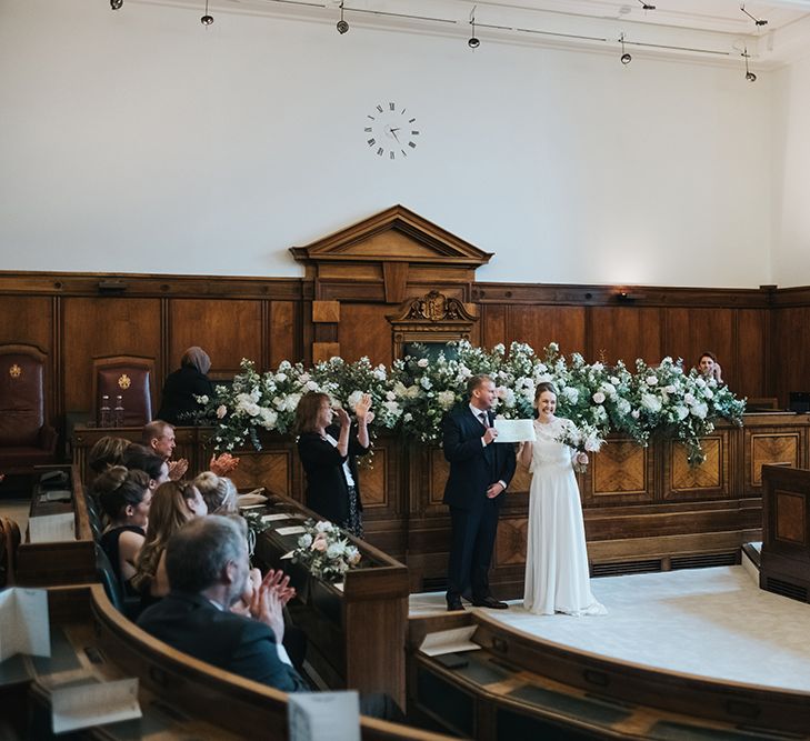 Blush and White Flowers with Foliage Altar Arrangement | Bride in Charlie Brear Carenne Dress with Spaghetti Straps and Flowing Skirt | Bridal Lace Jacket by Charlie Brear | Bouquet of White and Blush Flowers with Foliage | Groom in Navy Three-Piece Suit with Blush Pocket Square | Town Hall Hotel Wedding Ceremony | Statement Floral Arrangement Altar and Potted Plants with Copper Frame Table Plan and Lace Jacket | Miss Gen Photography