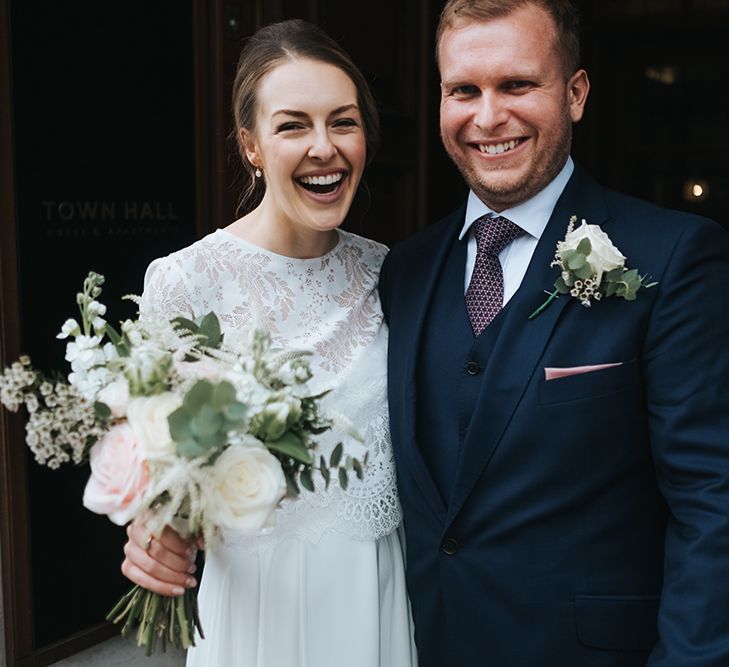 Bride in Charlie Brear Carenne Dress with Spaghetti Straps and Flowing Skirt | Bridal Lace Jacket by Charlie Brear | Bouquet of White and Blush Flowers with Foliage | Groom in Navy Three-Piece Suit with Blush Pocket Square | Statement Floral Arrangement Altar and Potted Plants with Copper Frame Table Plan and Lace Jacket | Miss Gen Photography