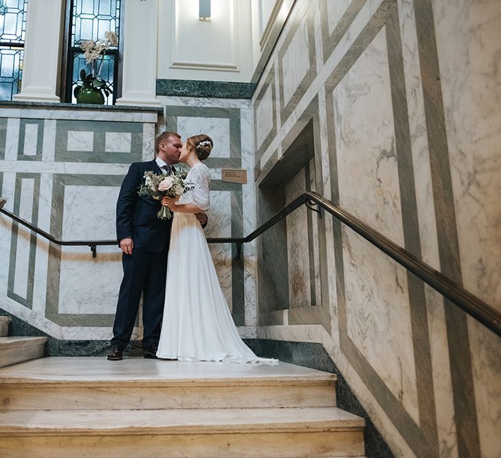 Bride in Charlie Brear Carenne Dress with Spaghetti Straps and Flowing Skirt | Bridal Lace Jacket by Charlie Brear | Porcelain Floral Hairpiece by Eden B | Bouquet of White and Blush Flowers with Foliage | Groom in Navy Three-Piece Suit with Blush Pocket Square | Hanging Lamps | Wedding Reception at Town Hall Hotel | Statement Floral Arrangement Altar and Potted Plants with Copper Frame Table Plan and Lace Jacket | Miss Gen Photography