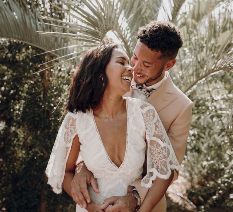 Groom in pale pink blazer embracing his boho bride in a Grace Loves Lace wedding dress